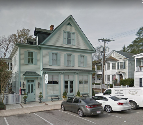 A light blue historic building with a peaked roof, surrounded by parked cars and trees, in a residential area.