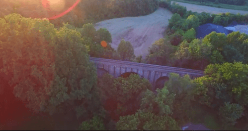 Aerial view of a stone bridge surrounded by lush green trees and a winding landscape under a bright sun.