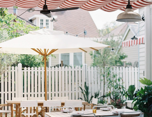 A cozy outdoor dining area with striped umbrellas, wooden tables, and a white picket fence surrounded by greenery.