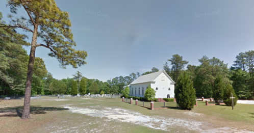A white church surrounded by trees, with a cemetery visible in the background on a sunny day.