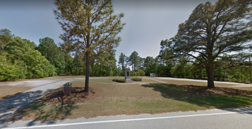 A quiet roadside scene featuring trees, a grassy area, and a monument surrounded by a paved road.