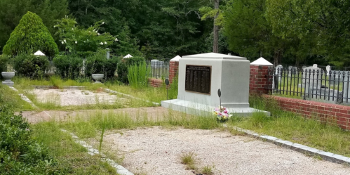 Overgrown cemetery with a monument and flower arrangement, surrounded by greenery and a wrought iron fence.