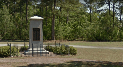 A tall monument surrounded by a black fence, set in a grassy area with trees in the background.
