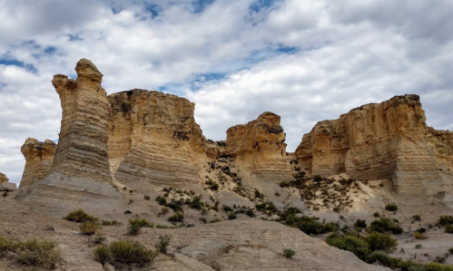 Rocky formations rise against a cloudy sky, showcasing layered textures and sparse vegetation in a desert landscape.