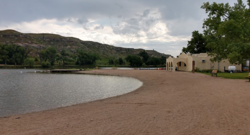 A sandy beach by a calm lake, with trees and a building in the background under a cloudy sky.