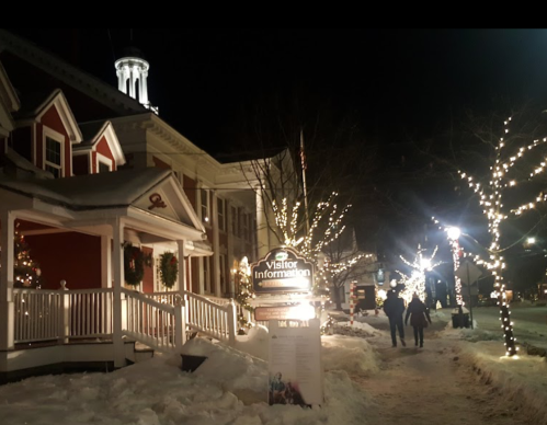 A snowy street at night, lined with festive lights and a visitor information sign, with people walking by.