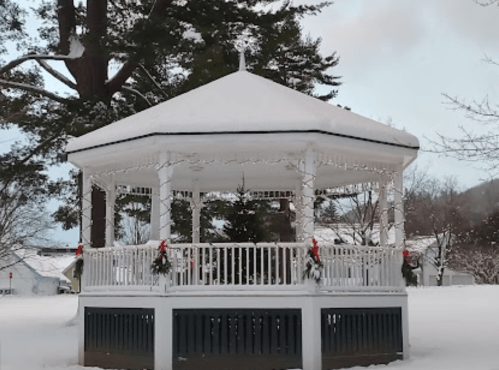 A snow-covered gazebo decorated with holiday garlands, surrounded by trees in a winter landscape.