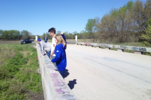A group of people stands on a bridge, looking over the edge at a grassy area below on a sunny day.