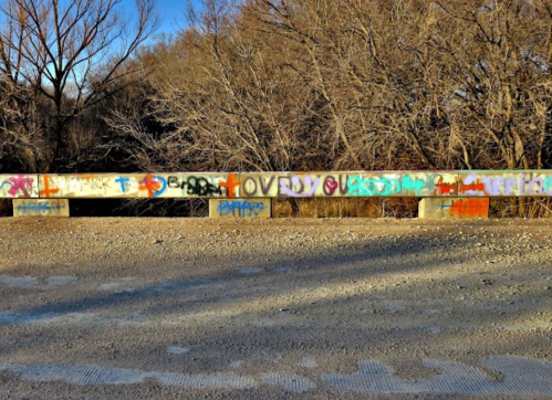 Colorful graffiti on a concrete barrier, surrounded by bare trees and gravel, under a clear blue sky.
