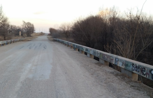 A gravel road with a bridge over a small creek, surrounded by bare trees and a cloudy sky.