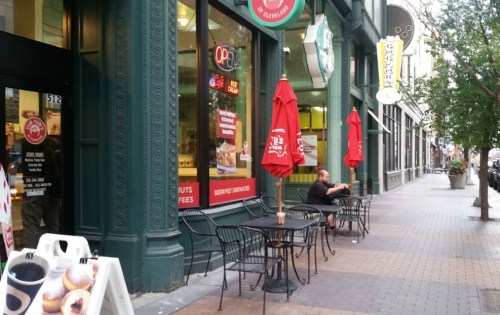 A sidewalk café with red umbrellas, a man sitting alone at a table, and a storefront in a city setting.