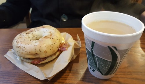 A bagel sandwich with bacon on a paper wrapper next to a cup of tea on a wooden table.