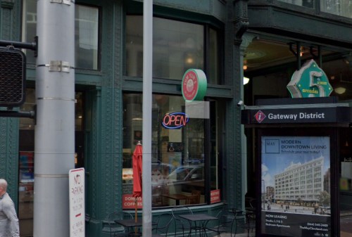 A storefront with an "OPEN" sign, featuring outdoor seating and a nearby bus stop in an urban setting.