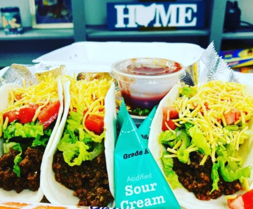 Three tacos filled with seasoned meat, lettuce, tomatoes, and cheese, served with sour cream and salsa on the side.