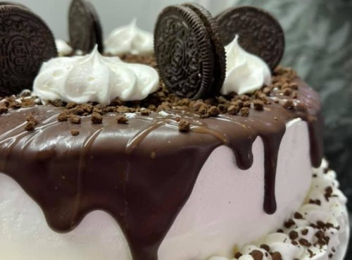 A chocolate cake topped with whipped cream, crushed cookies, and whole Oreo cookies, with chocolate drizzle.