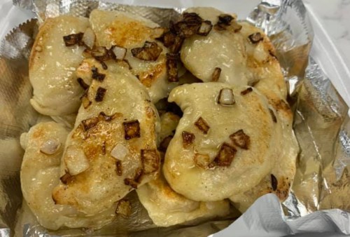 A tray of golden-brown pierogi topped with sautéed onions, served in a foil container.