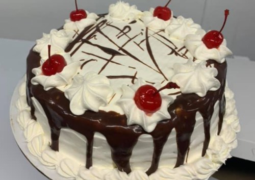 A round chocolate cake topped with whipped cream, chocolate drizzle, and cherries.
