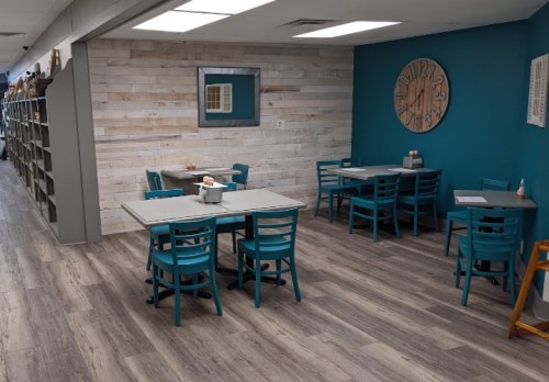 A cozy dining area with teal chairs and tables, wooden walls, and a large clock, creating a welcoming atmosphere.