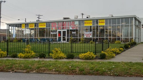 A grocery store with a deli and market, featuring large windows and a green lawn in front. Signs indicate it's open.