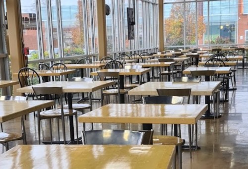 Empty dining area with wooden tables and black chairs, surrounded by large windows letting in natural light.