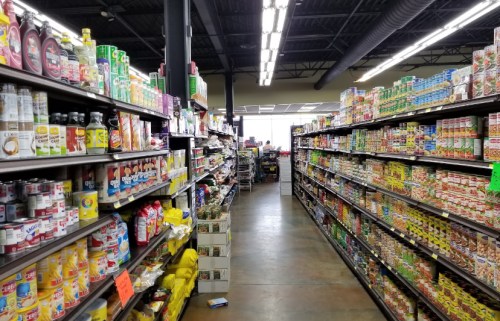 A grocery store aisle filled with various canned goods, snacks, and beverages on both sides. Brightly lit interior.