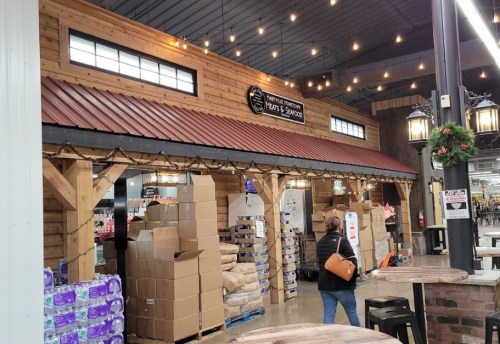 Interior of a market with wooden structures, stacked boxes, and a person walking by under string lights.
