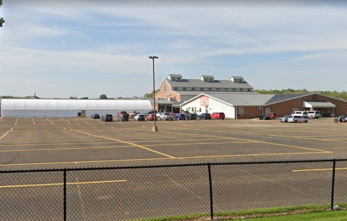 A large building with a white tent structure nearby, surrounded by a spacious parking lot and a fence in the foreground.