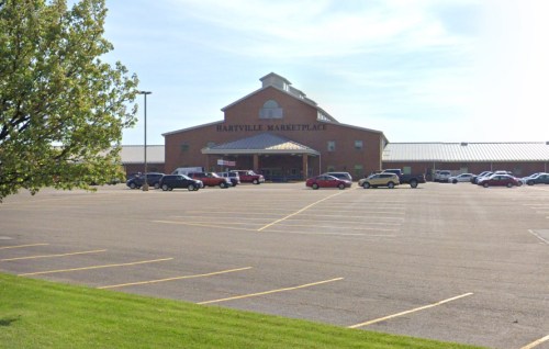 Hartville Marketplace building with a large parking lot, surrounded by grass and trees under a clear blue sky.