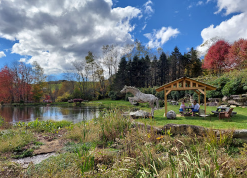 A serene landscape featuring a pond, colorful trees, and a gazebo with people enjoying the outdoors.