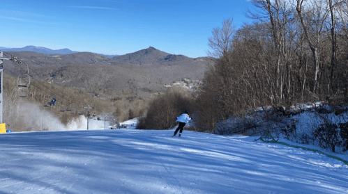 A skier glides down a snowy slope with mountains in the background under a clear blue sky.