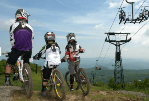 Three mountain bikers in gear overlook a scenic view, with a ski lift in the background on a sunny day.