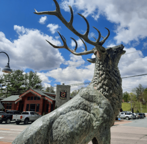 A large bronze statue of a deer with antlers, set against a blue sky with clouds and a building in the background.