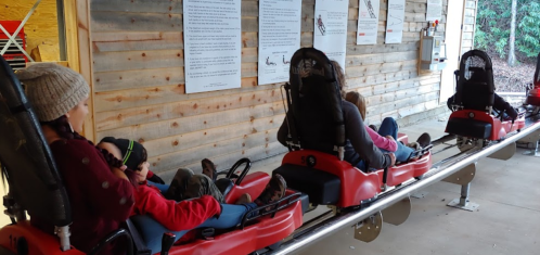 A group of people seated in red sleds, preparing for a ride at an outdoor attraction, with informational signs in the background.