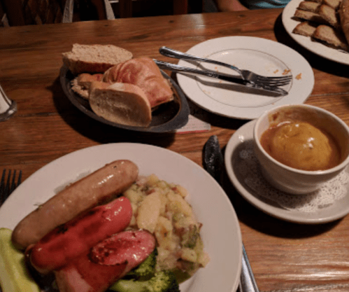 A plate with sausage, vegetables, and potatoes, alongside bread and a bowl of soup on a wooden table.