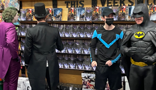 Four people in costumes, including Batman and the Joker, stand in a comic book store with shelves of comics behind them.