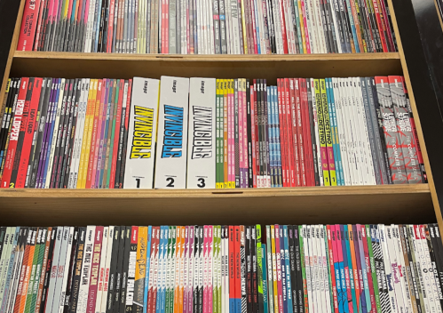 A wooden shelf filled with various comic books and graphic novels, organized by series and color.