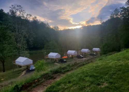 A serene landscape featuring white cabins by a river at sunset, surrounded by trees and grassy hills.