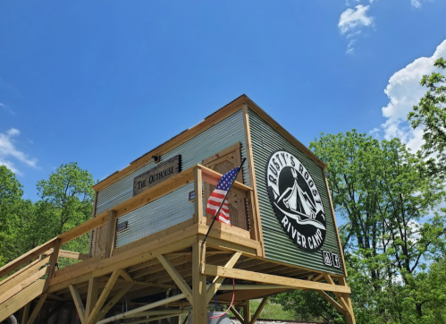 A wooden structure with a sign reading "Rusty's River Cabin" and an American flag, set against a blue sky and green trees.