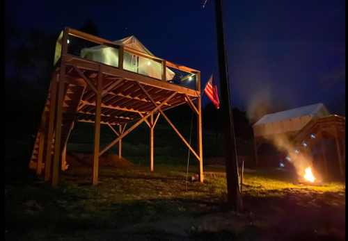 A wooden structure elevated at night, with an American flag and a fire burning nearby.