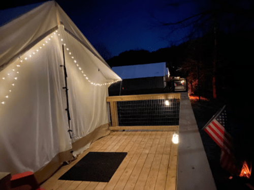A cozy outdoor scene at night featuring a tent with string lights, a wooden deck, and an American flag.