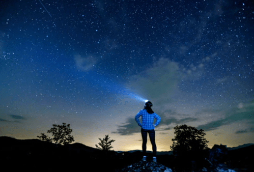 A person stands on a rock, gazing at a starry sky, with a headlamp illuminating the surroundings.