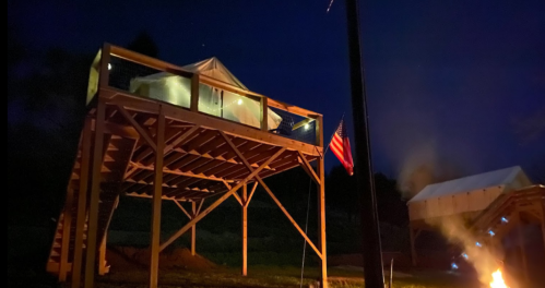 A wooden elevated structure at night, featuring a tent and an American flag, with a fire nearby.