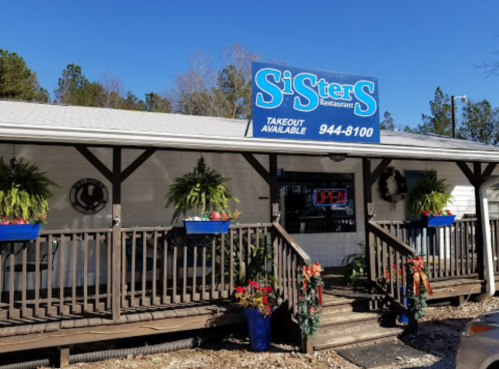 A cozy restaurant exterior with a sign reading "Sisters Restaurant," featuring a porch and colorful flower pots.