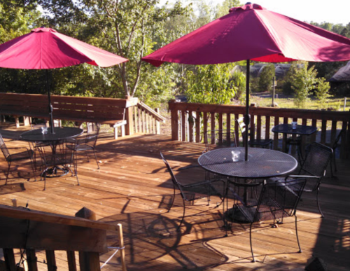 A wooden deck with black tables and chairs, shaded by red umbrellas, surrounded by greenery.