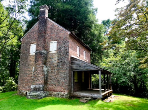 A historic brick house with boarded windows, surrounded by lush greenery and trees.