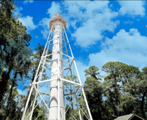 A tall white lighthouse tower surrounded by trees under a bright blue sky with scattered clouds.