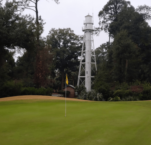 A golf course with a flag on the green and a tall lighthouse in the background, surrounded by trees.