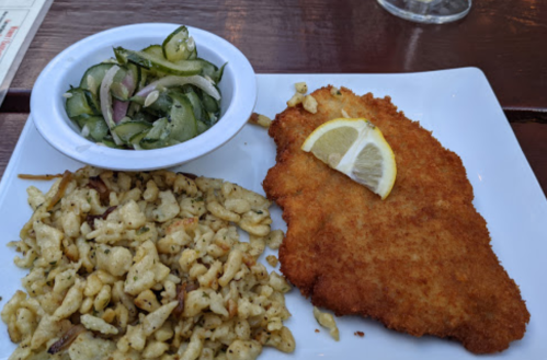 A plate featuring a golden-brown breaded fish fillet with a lemon slice, served alongside cucumber salad and pasta.