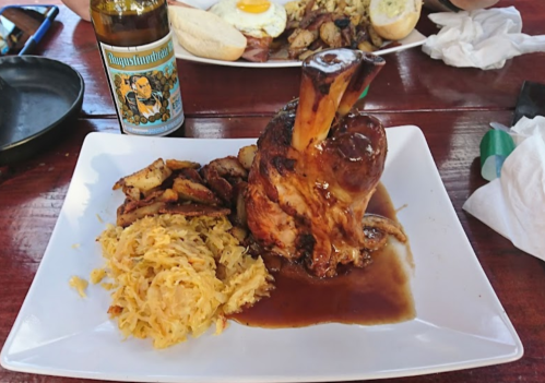 A plate featuring a large pork knuckle with gravy, sauerkraut, and roasted potatoes, alongside a bottle of beer.