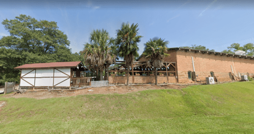 A brick building with a red roof, surrounded by palm trees and a grassy area, featuring a wooden deck and outdoor decor.